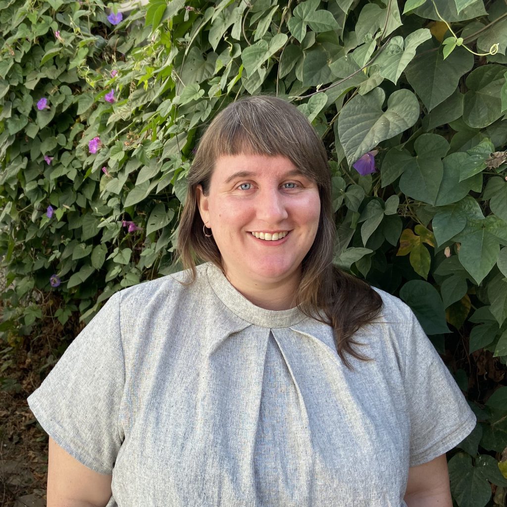 fat, white woman with blue eyes and light brown hair, smiling and standing in front of a wall of leaves, wearing a gray linen shirt with pleats at the neckline
