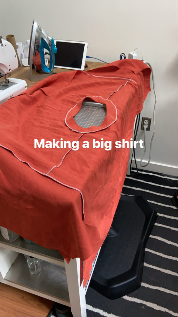 White text reads "making a big shirt" on top of a spread open red fabric that covers an entire table top. There are constrasting white serged edges around all of the sewn seams.