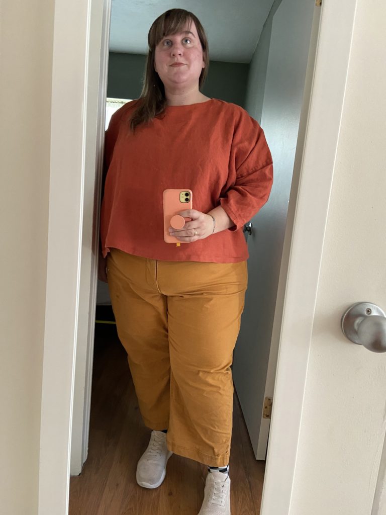Traci, a fat white woman stands in front of a full length mirror wearing a slightly oversized boxy red linen shirt with tumeric yellow wide leg pants and creamy tan sneakers